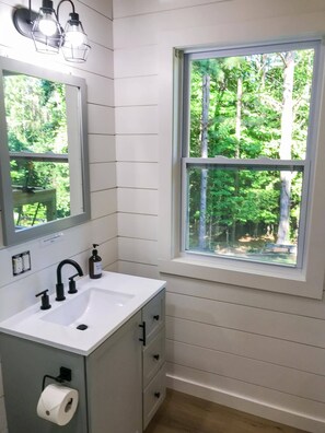 A bathroom with a white sink, black faucet, and a mirror above it. There is a window with a view of trees outside. - Cozy Clemson TIny House Cabin (Seneca)
