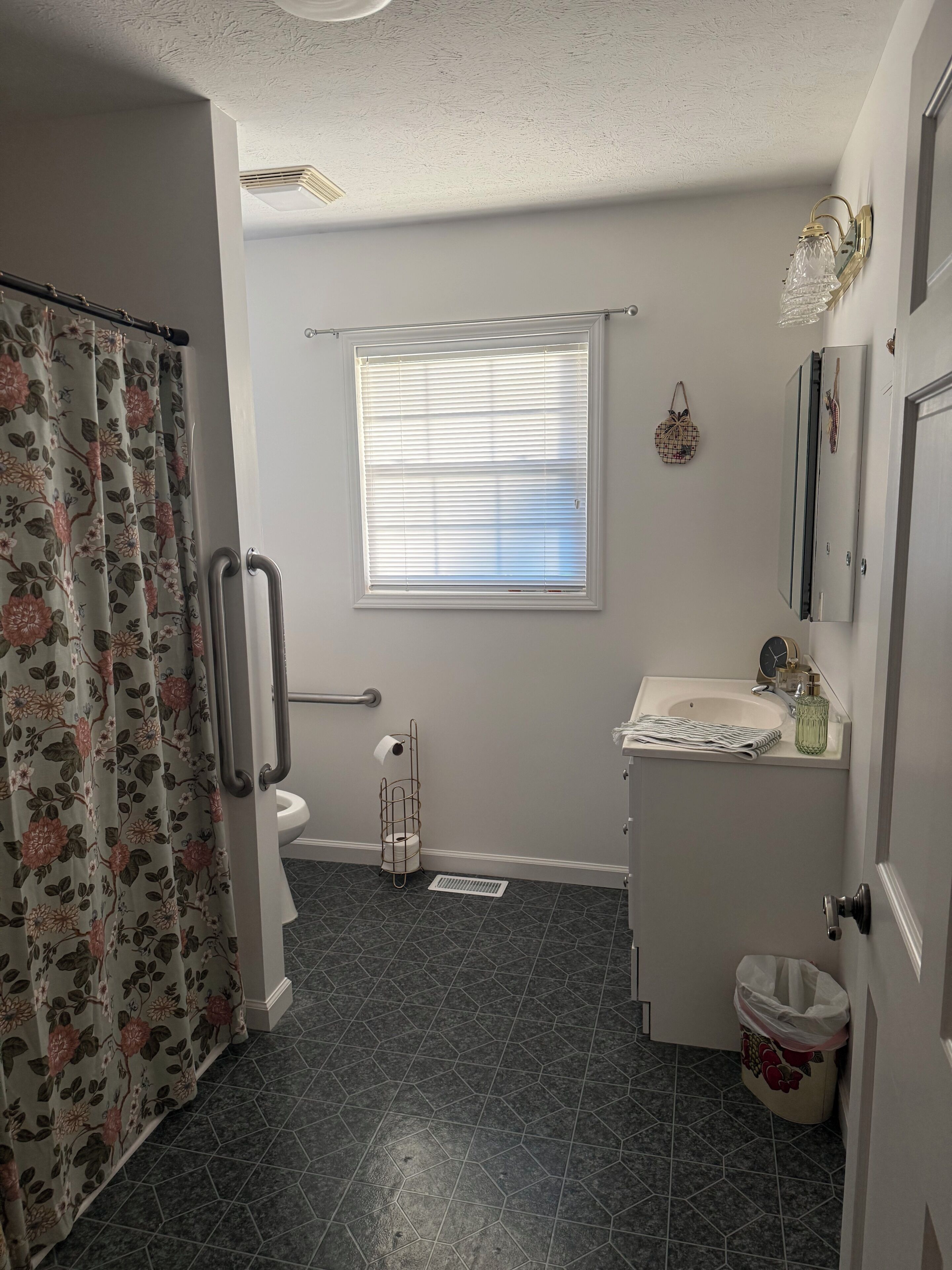 A bathroom with a shower curtain, toilet, window with blinds, sink, and a trash can.