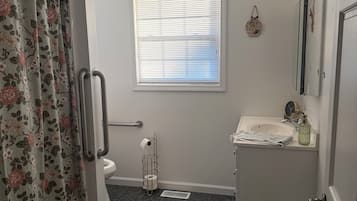 A bathroom with a shower curtain, toilet, window with blinds, sink, and a trash can.