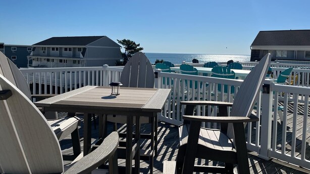 A wooden deck with tables and chairs, overlooking a beach with buildings in the background. - Beachwalk 20, Steps to the Beach, Outdoor Pool (Ocean City)