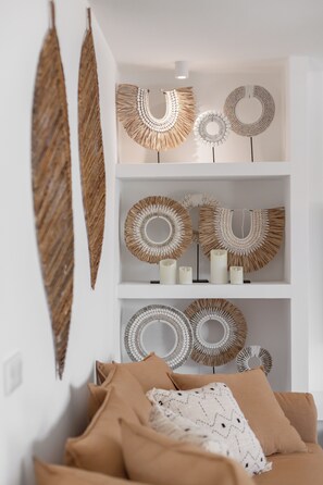Ein Zimmer mit einer weißen Wand, einer hängenden Blattschmuckdeko und einem Sofa mit beige farbenen Kissen.