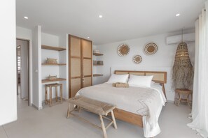 A bedroom with a wooden bed, two bedside tables, a bench, a wardrobe, and wall decor. - Villa Kaia (oyster pond)