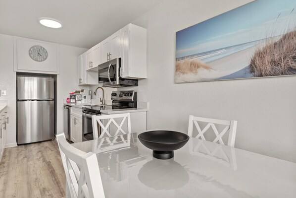 A modern kitchen with white cabinetry, stainless steel appliances, and a dining area with a black bowl on the table. - Tropical Isle 209🌴Beach Access👙 (Fort Walton Beach)