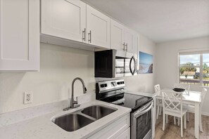 A kitchen with white cabinets, a stainless steel sink, a stove, and a dining area with white chairs. - Tropical Isle 209🌴Beach Access👙 (Fort Walton Beach)