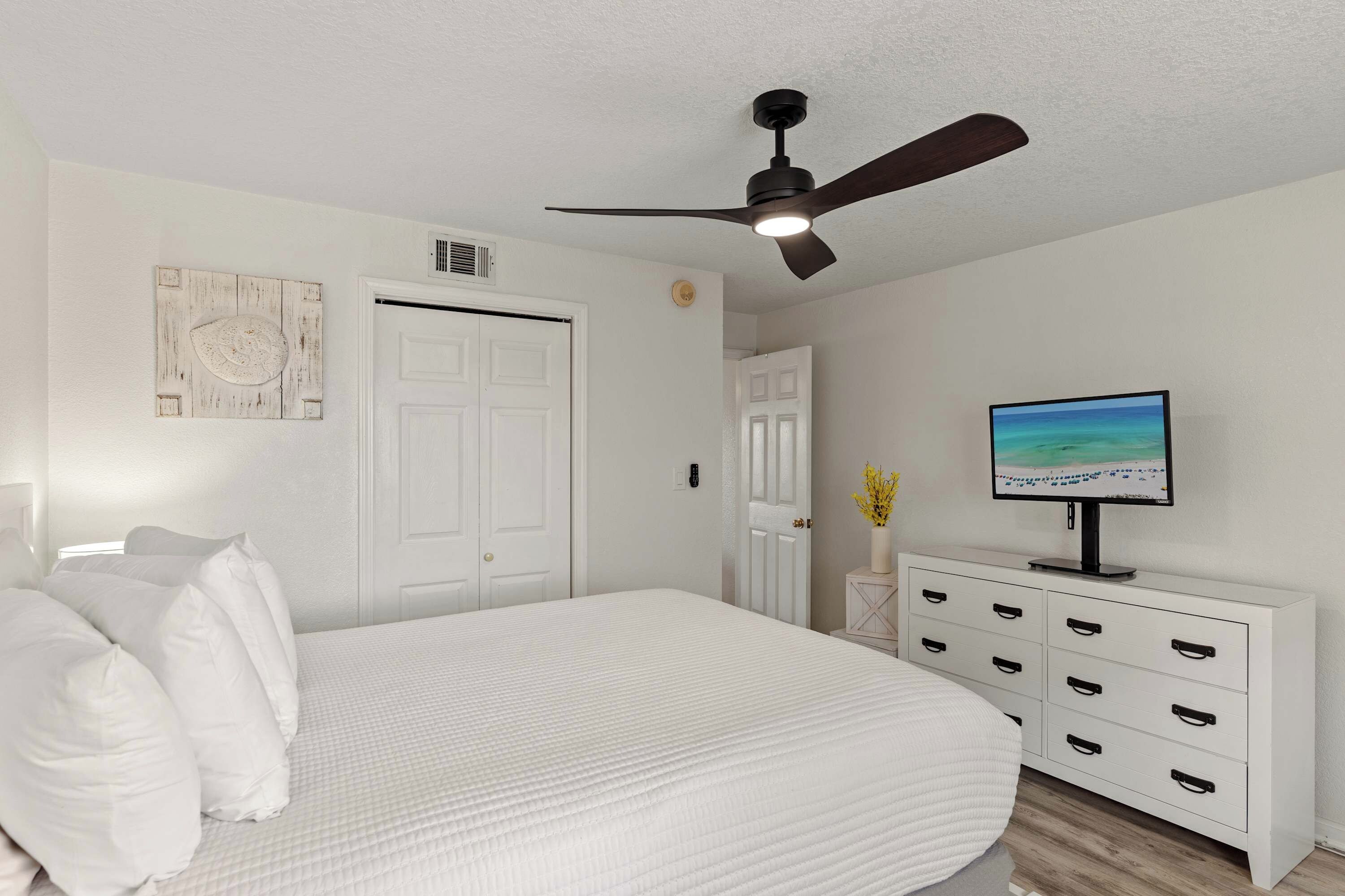 A bedroom with a bed, a ceiling fan, a white dresser, a television, and a wall-mounted artwork.