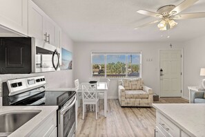 A modern kitchen with white cabinetry, a built-in oven, and a microwave. There is a dining area with a white table and chairs, and a view of palm trees through a large window. - Tropical Isle 209🌴Beach Access👙 (Fort Walton Beach)
