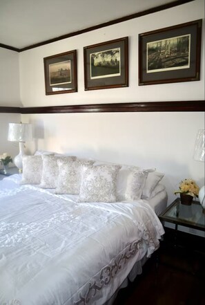 Een net opgemaakt bed met een witte spreideken en bijpassende kussens, een nachtkastje met een lamp, en drie ingelijste foto’s aan de muur boven het bed.