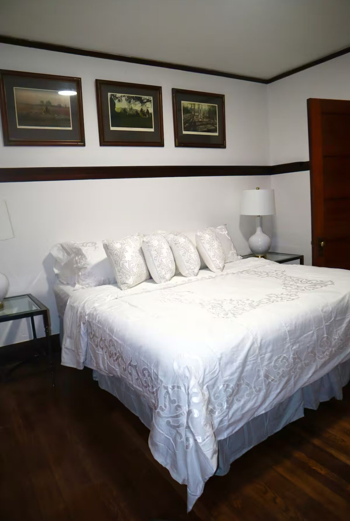 A neatly made bed with white linens and decorative pillows, a bedside lamp, and framed pictures on the wall.