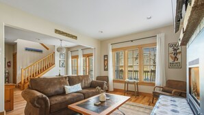 A spacious living room with a brown leather sofa, a wooden coffee table, and a large window with blinds. A staircase is visible in the background leading to another level. - Pine Haven Retreat | Walk to Downtown & Pool (Sisters)