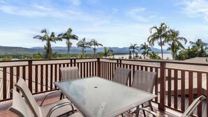 A deck with a glass-top table and chairs, overlooking a beach with palm trees and mountains. - Vistas @ The Point Su (Port Douglas)