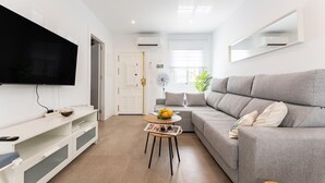 Ett modernt vardagsrum med en platt-TV, en grå soffa, en vit TV-bänk och ett litet bord med en tidning och en fruktskål.