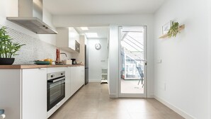 Ett modernt kök med vita skåp, en inbyggd ugn och en köksfläkt i rostfritt stål. På bänkskivan står en växt och genom en öppen dörr kan man se ut mot omgivningen.