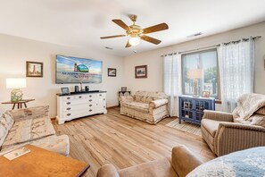 Ein Wohnzimmer mit Deckenventilator, Fernseher, Sofa, Couchtisch und einem Fenster mit Vorhängen.