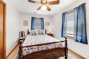 A bedroom with a wooden bed, a ceiling fan, two windows with blue curtains, and a nightstand with a lamp. - Bradford House by Seaside Vacations & Sales (Chincoteague)