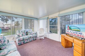 A room with a floral sofa, a white chair, a desk, and a large window with a view of the outdoors. - Bradford House by Seaside Vacations & Sales (Chincoteague)