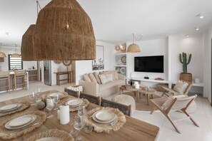 Ein modernes Wohnzimmer mit Essbereich, ausgestattet mit einer großen, gewebten Pendelleuchte, einem Flachbildfernseher und einer Kakteenpflanze.