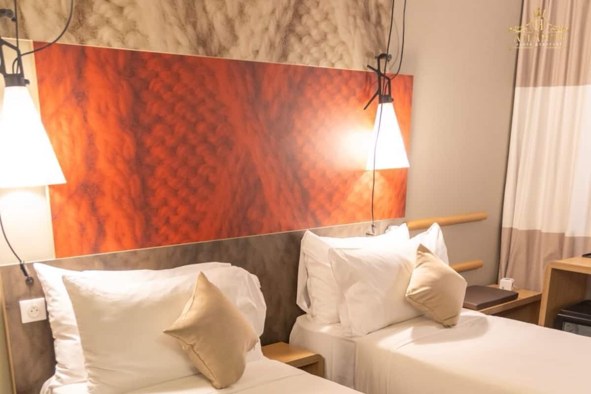 Ein Hotelzimmer mit zwei Einzelbetten, beide mit weißen Laken und beigen Kissen. Am Kopfteil befindet sich ein Holzpaneel mit einem strukturierten, orangefarbenen Muster, und es gibt zwei Nachttischlampen. Auf der rechten Seite ist ein kleiner Nachttisch mit einer Lampe zu sehen.