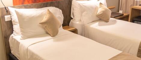 A hotel room with two single beds, each with white linens and beige pillows. There is a wooden headboard with a textured orange design, and two bedside lamps. A small wooden nightstand with a lamp is visible on the right side.