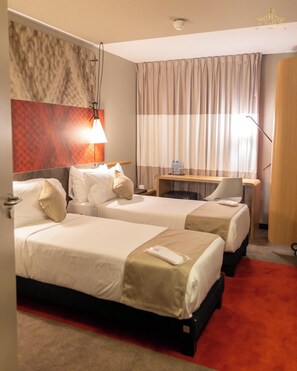 A hotel room with two beds, a desk, a chair, a lamp, and a large window with curtains. - Atlantis Alger Aéroport ex Ibis (Algiers)