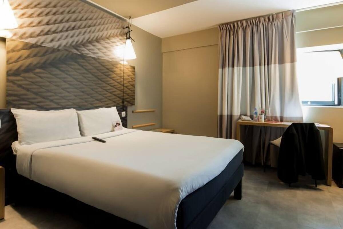 Ein Hotelzimmer mit Bett, Nachttisch, Lampe und Blick durch ein Fenster nach draußen.