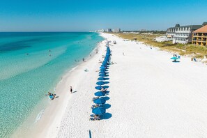 Ein Strand mit einer Reihe blauer Sonnenschirme und Liegestühlen, einem weißen Sandstrand und Gebäuden im Hintergrund.