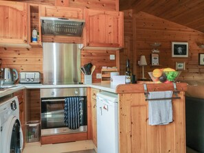 A compact kitchen with wooden cabinetry, a stainless steel range, a washing machine, and a small refrigerator. - Watendlath (Keswick)