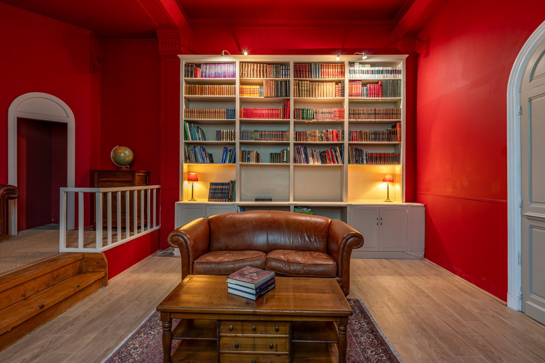 Une pièce avec un mur rouge, un canapé en cuir, une table basse en bois et une grande bibliothèque remplie de livres.