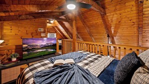 A cozy log cabin bedroom with a bed, a television displaying a scenic landscape, and a ceiling fan. - Hot Tub, View, Pool Table, Fireplace, Honeymoon! (Sevierville)