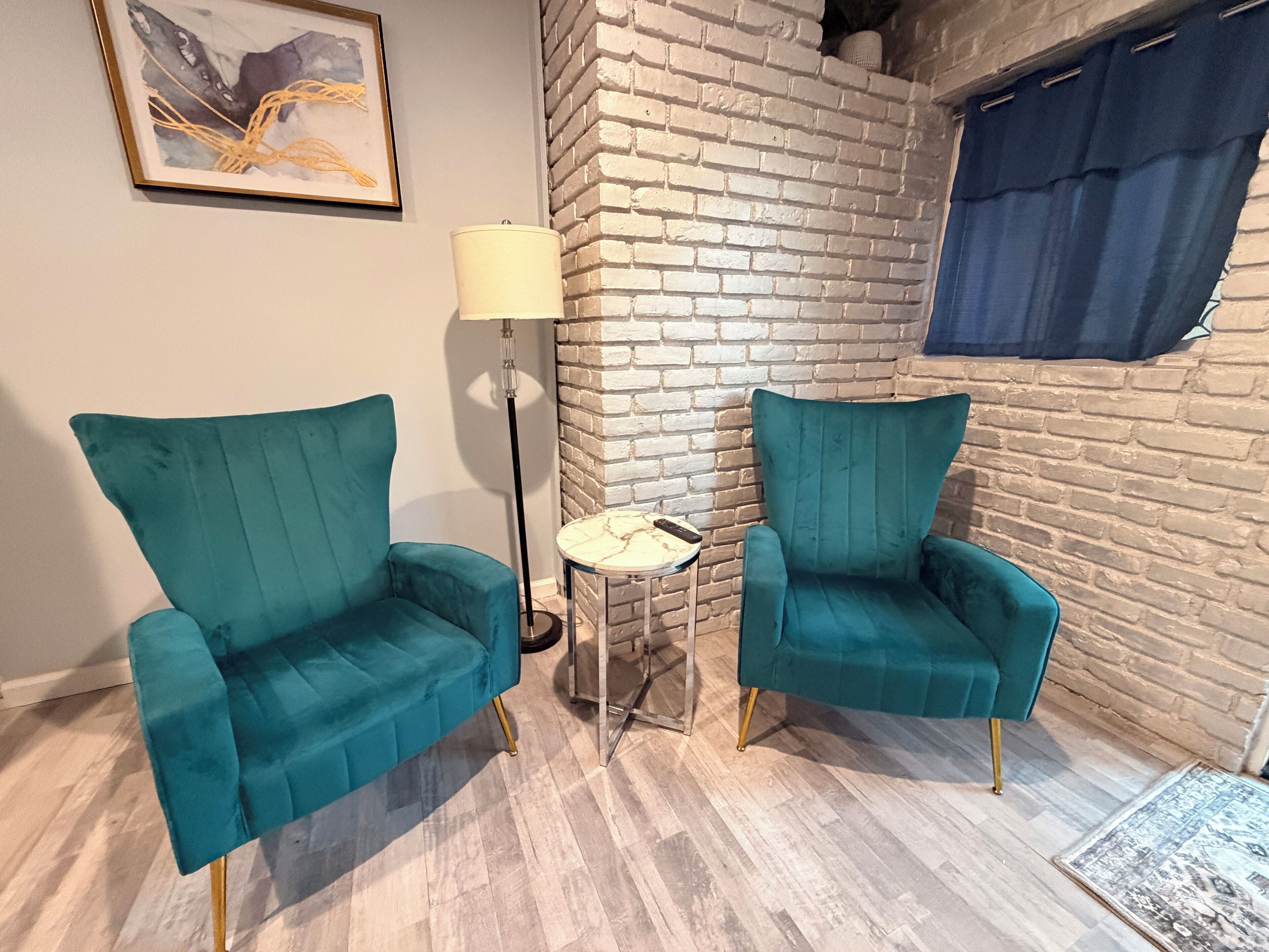 Ein Zimmer mit zwei türkisfarbenen Sesseln, einer Stehlampe, einem kleinen runden Tisch und einer Ziegelwand.
