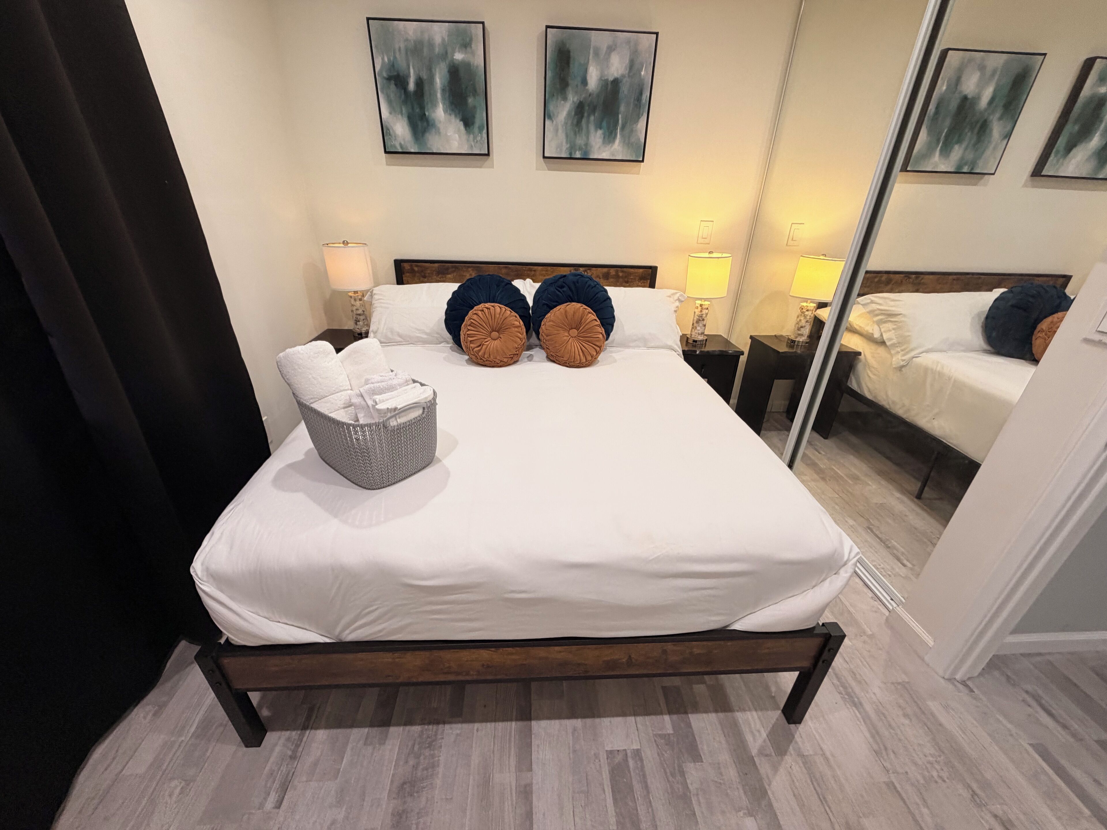 Un lit bien fait, avec du linge de lit blanc, deux tables de chevet avec des lampes, un panier en osier gris et des tableaux abstraits accrochés au mur.
