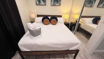 Un lit bien fait, avec du linge de lit blanc, deux tables de chevet avec des lampes, un panier en osier gris et des tableaux abstraits accrochés au mur.