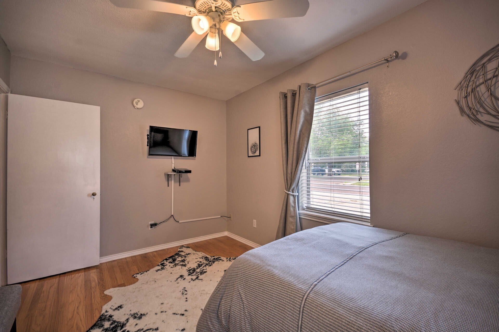 A bedroom with a bed, a ceiling fan, a TV mounted on the wall, a window with blinds, and a door.