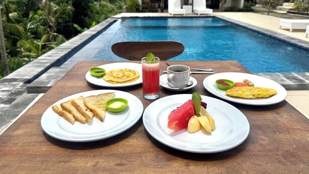 A breakfast setting by a pool with plates of toast, pancakes, and fruit, and a drink. - The Lebah Kelingking Villa (Penida Island)
