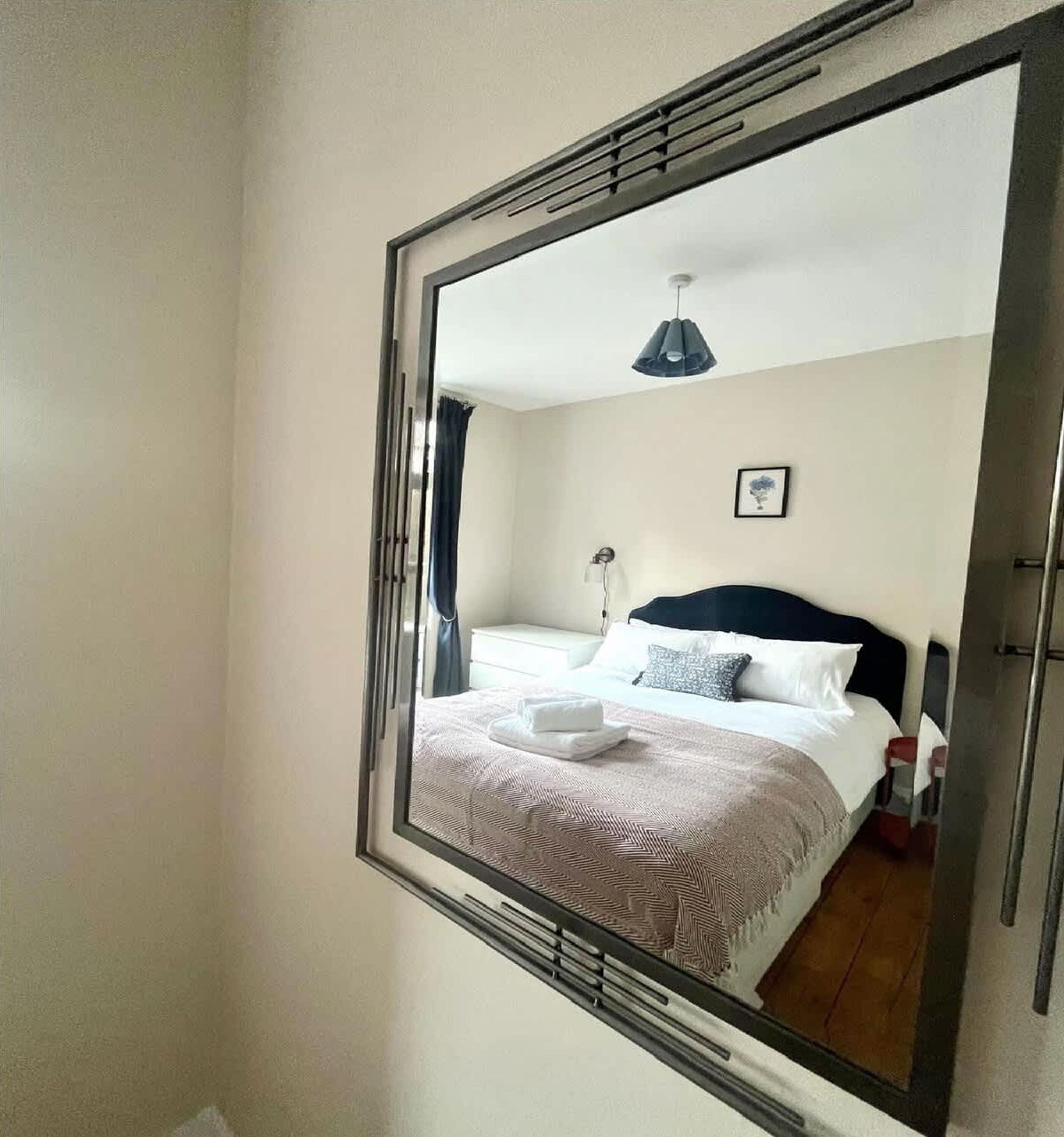 A hotel room with a large mirror, a bed with a white headboard, a bedside table, a lamp, and a picture on the wall.