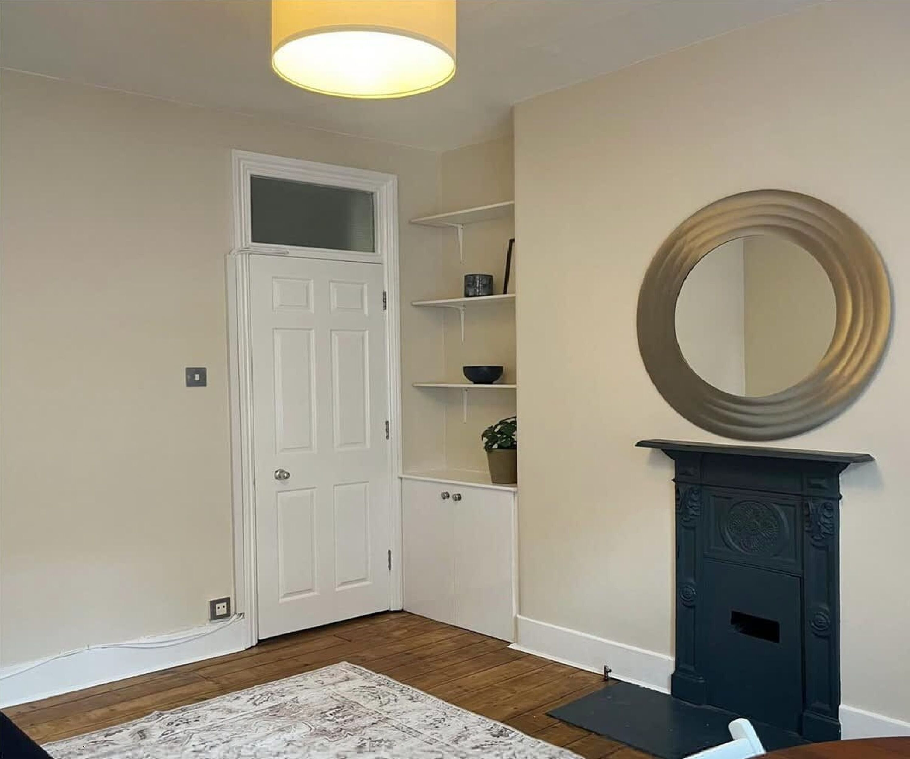 Un couloir avec une porte blanche, un plancher en bois, un tapis blanc, une cheminée surmontée d’un miroir et une étagère avec une plante.