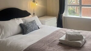Een hotelkamer met een bed, nachtkastje, een wandlamp, een raam met gordijnen en een lijst met een afbeelding aan de muur.