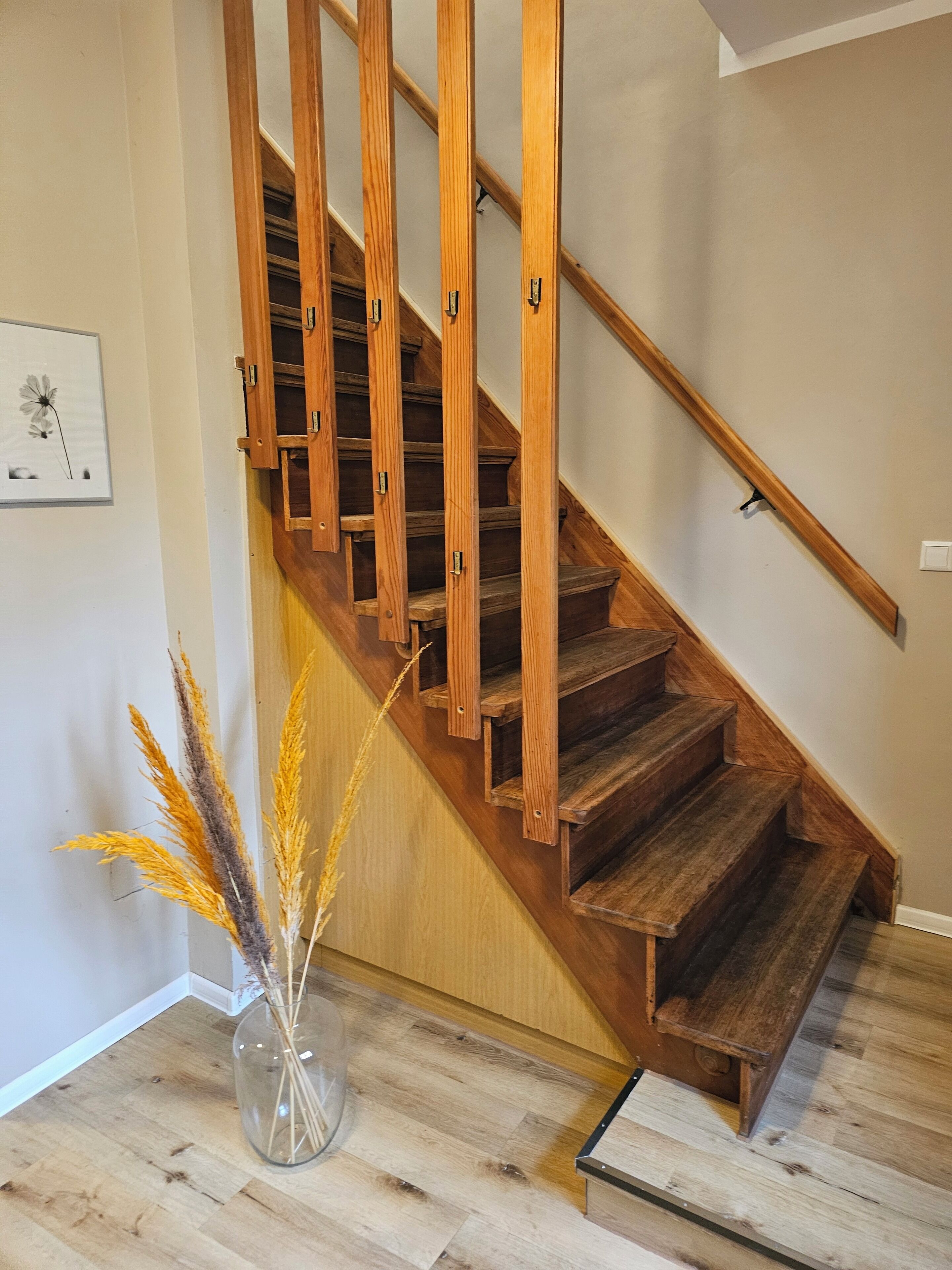 Una escalera de madera con barandilla, un jarrón de vidrio transparente con plantas secas en el suelo y un cuadro en la pared.