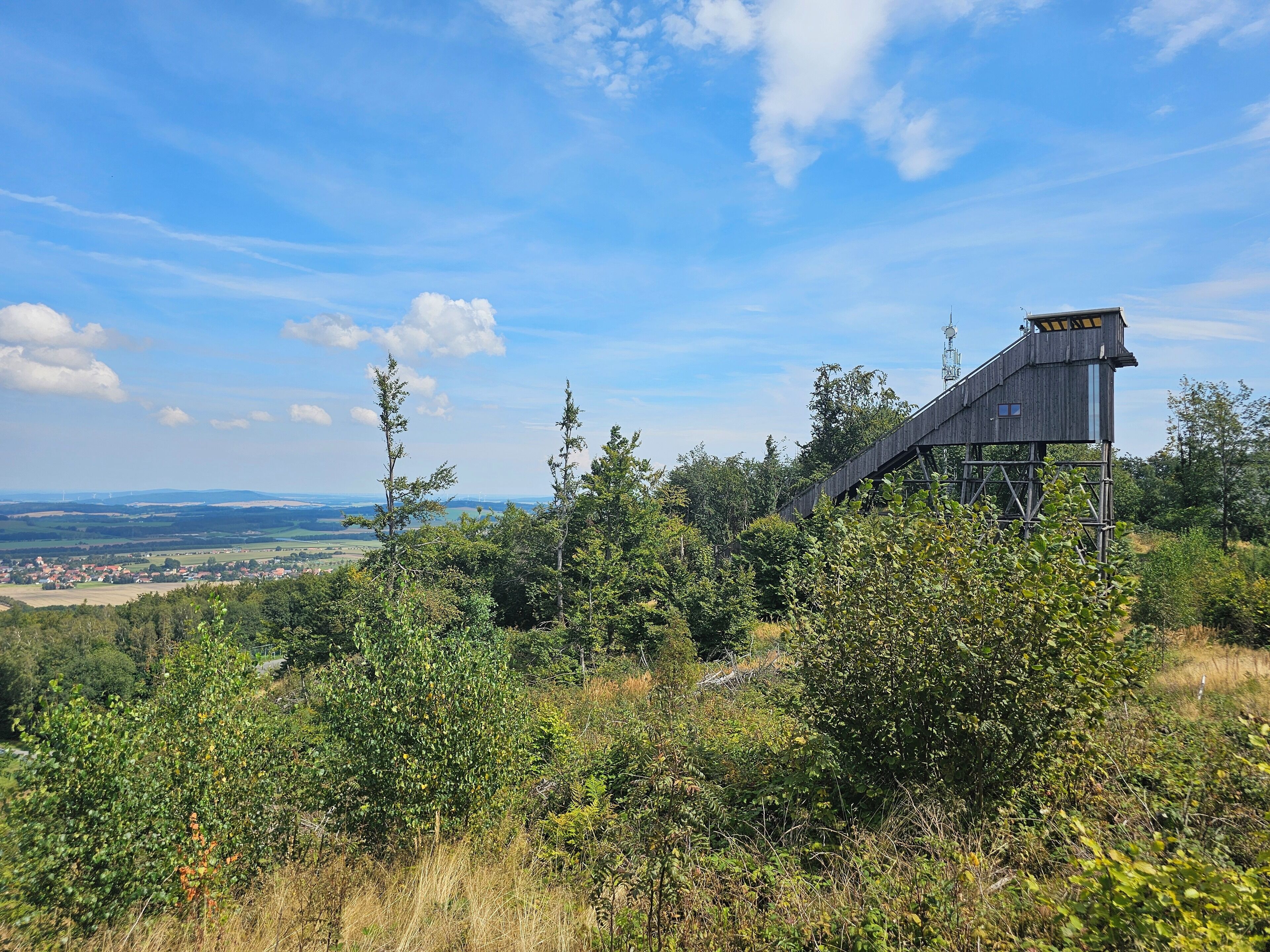 A wooden observation tower with a staircase leading up to it, surrounded by dense greenery and overlooking a distant town.