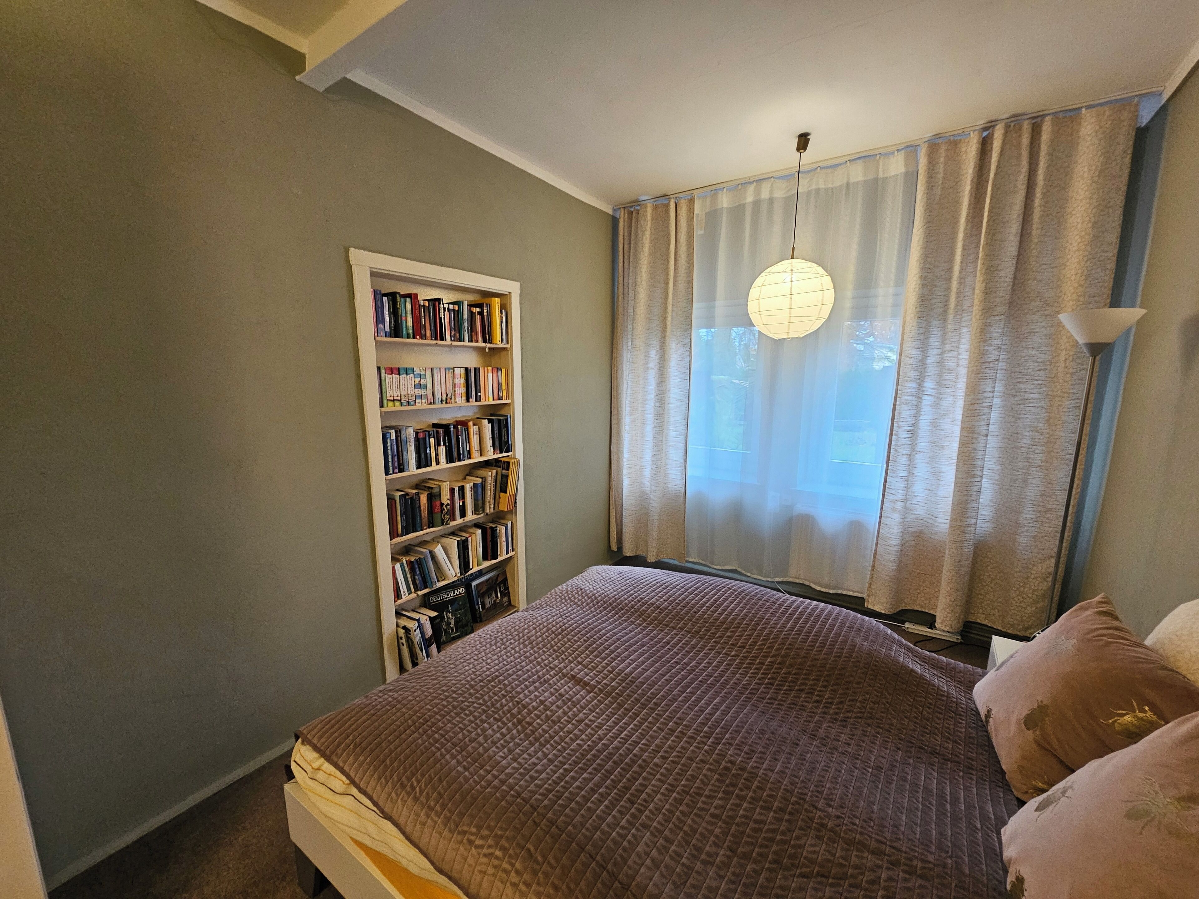 Un dormitorio con cama, estantería, ventana con cortinas y lámpara colgante.