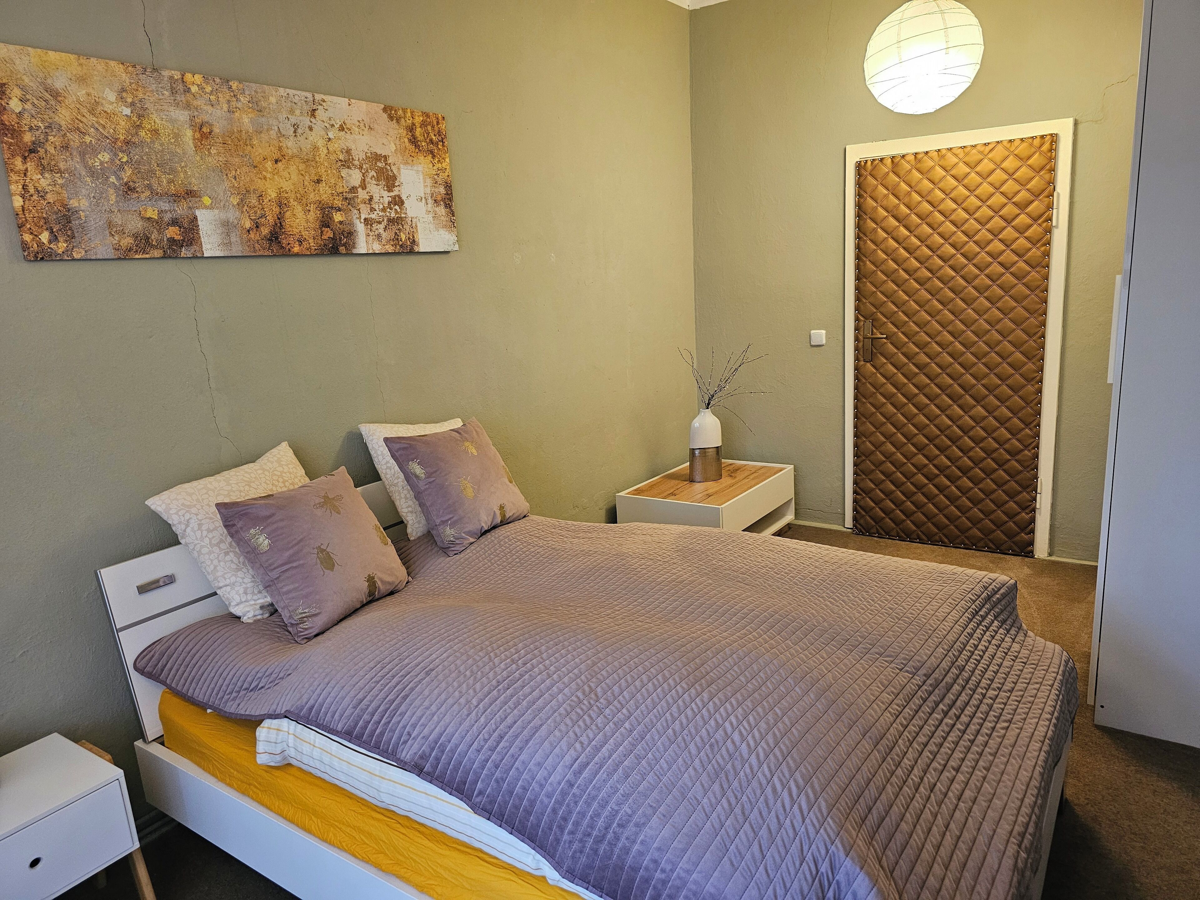 Ein Schlafzimmer mit einem Bett, Nachttischen, einem Beistelltisch, einem Wandbild und einer Tür mit einer gesteppten Zarge.