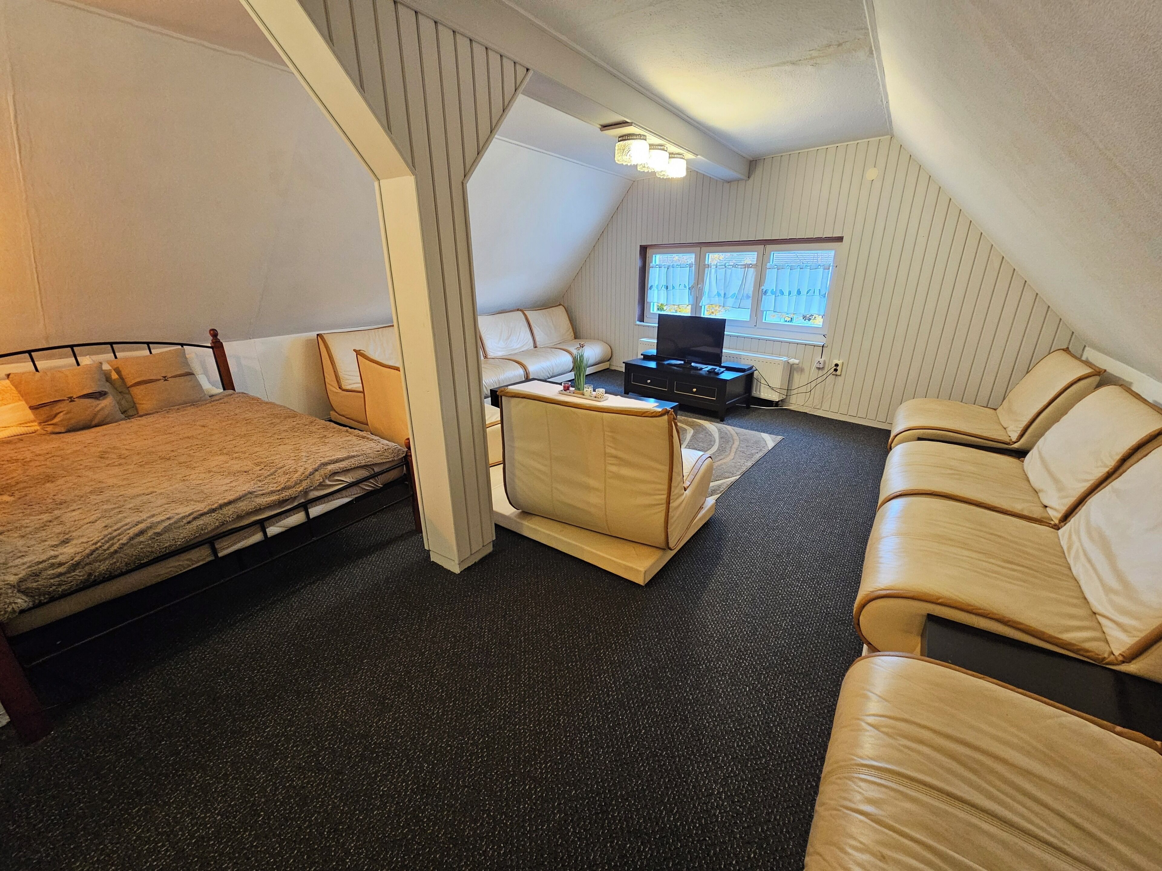 Ein kleiner, gut beleuchteter Raum mit einem Bett, einer Schlafcouch und einem kleinen Tisch. Ein Fernseher steht auf einem Schrank, und es gibt ein Fenster mit Vorhängen.