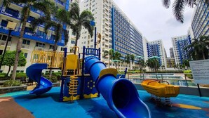 A playground with a blue slide and yellow play structures, surrounded by tall buildings and palm trees. - Stylish 1-Bedroom at Sea Residences, Pasay — close to SM Mall of Asia! (Pasay City)