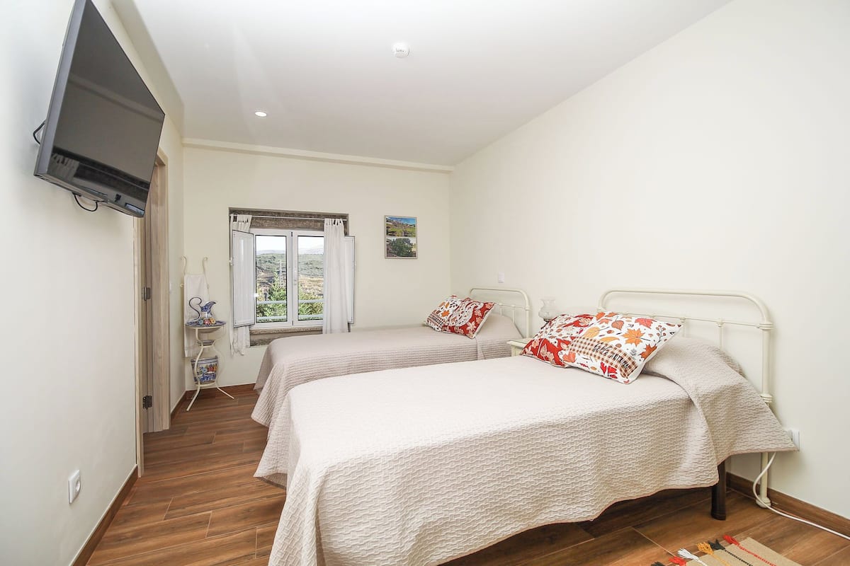 A bedroom with two beds, a TV mounted on the wall, a window with curtains, and a small table with a vase.