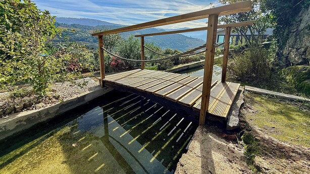 A wooden bridge over a clear water channel, surrounded by greenery and hills in the background. - Bosque da Fantasia (Baiao)
