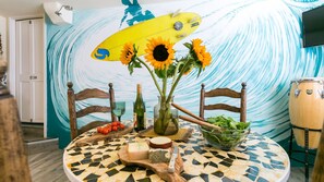 A dining table with a mosaic tile top, surrounded by wooden chairs. A surfboard, sunflowers, and a bowl of salad are on the table.