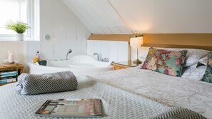 A bedroom with a large bed, a bedside table with a lamp, a window with a plant, and a bathroom with a bathtub and shower.