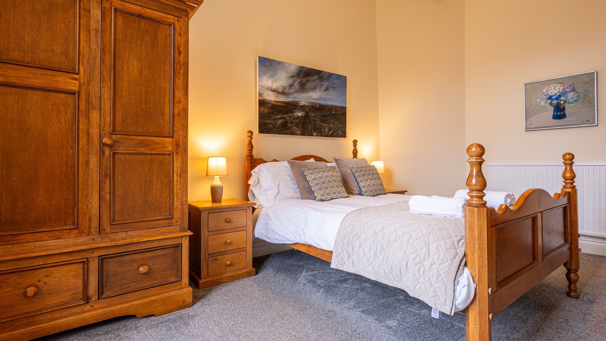 A bedroom with a large wooden wardrobe, a bed with a white duvet, patterned pillows, and a nightstand with a lamp.