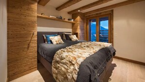 Ein Schlafzimmer mit Holzbalkendecke, einem Bett mit Felldecke, einem Fenster mit Bergblick und einem Regal mit Büchern und einer Lampe.