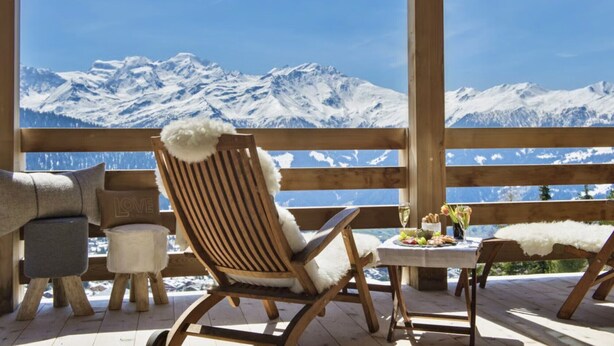 Eine Holzterrasse mit einem Holzstuhl und einem Hocker, ein kleiner Tisch mit einer Vase und Speisen, und Blick auf schneebedeckte Berge.