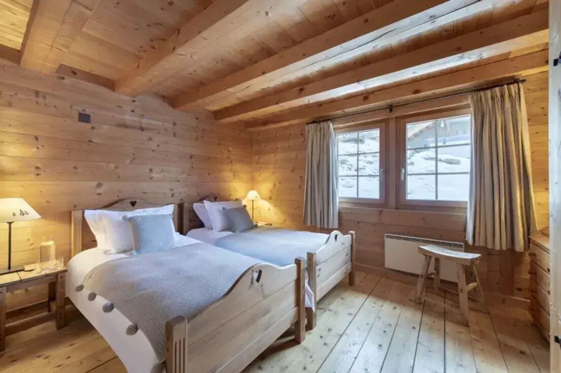 Une pièce chaleureuse, aménagée dans un chalet en bois, avec deux lits, une petite table et une fenêtre avec des rideaux.